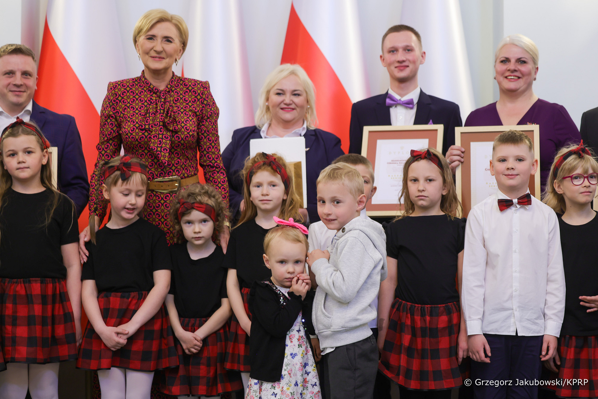 Osoby ustawione w dwóch rzędach. W pierwszym dzieci, w drugim dwóch mężczyzn i trzy kobiety. Dwie kobiety i dwóch mężczyzn trzymają w rękach dyplomy w ramach. Wszyscy elegancko ubrani. Z tyłu biało-czerwone flagi. 