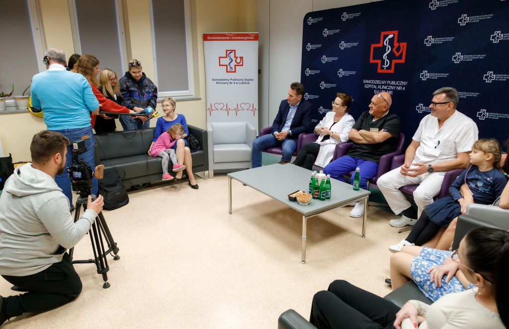 Spotkanie prasowe na temat żywienia pozajelitowego w ciąży. Z lewej strony dziennikarze z kamerą i mikrofonami rozmawiającymi z pacjentką i jej córką. Po prawej stronie na kanapach siedzą specjaliści szpitala i mamy z dziećmi.