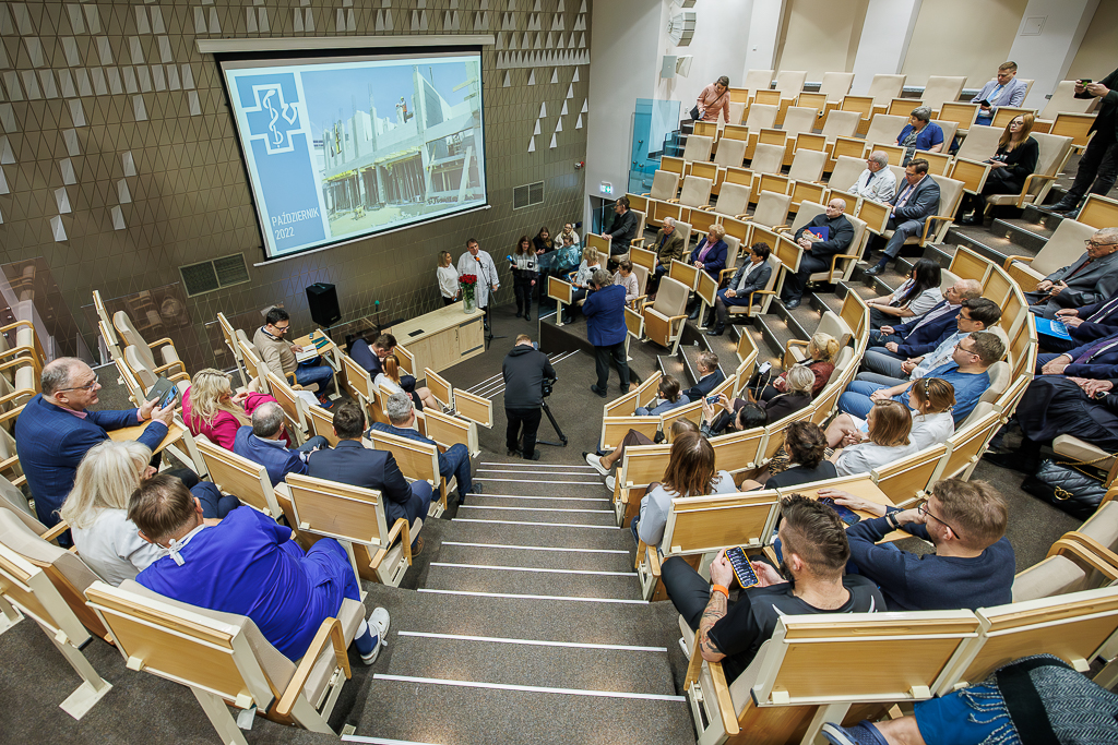 Kaskadowa aula wypełniona w połowie ludźmi. Na ekranie z tyłu wyświetlone zdjęcie z budowy budynku Stacji Hemodializ z napisem "Październik 2022".