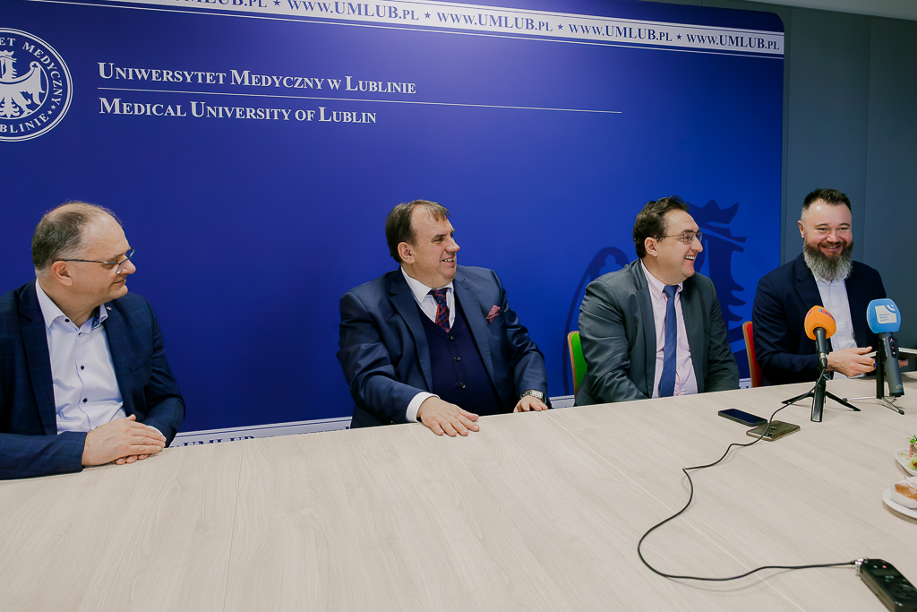Czterech uśmiechniętych mężczyzn w garniturach siedzących za stołem, na którym są mikrofony dziennikarskie