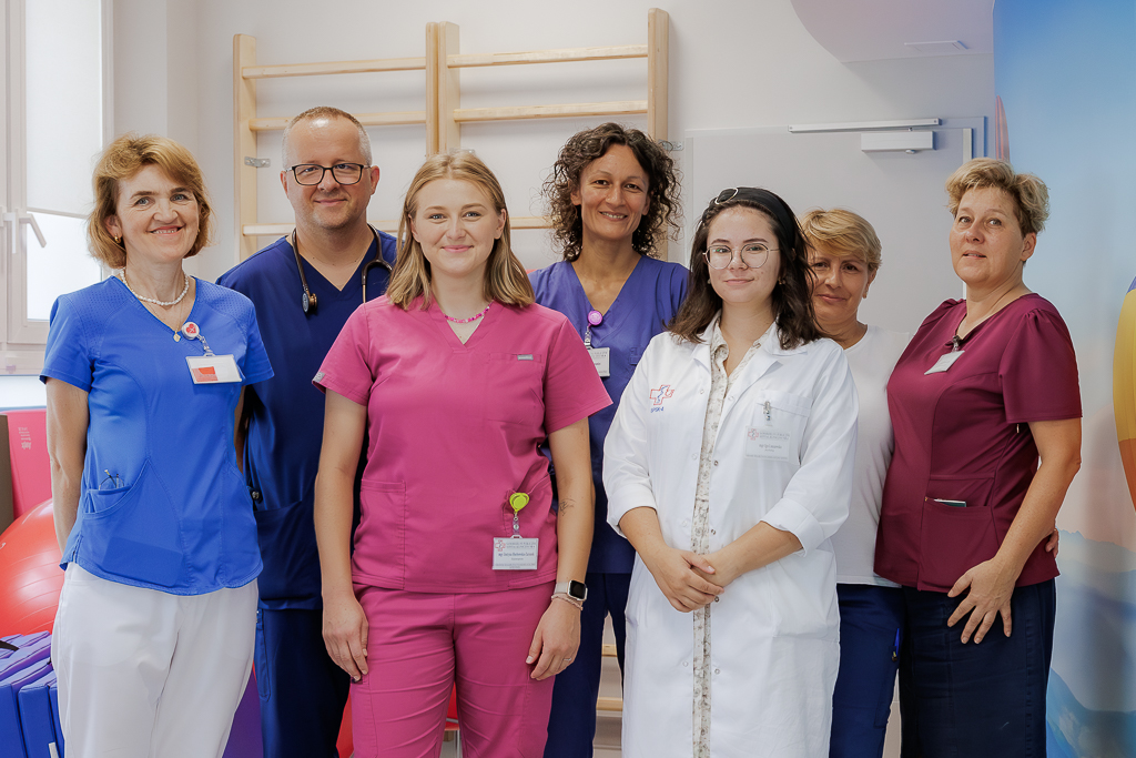 Siedem osób pracujących w ośrodku - lekarze, fizjoterapeutki, psycholog i pielęgniarka, stoją obok siebie na tle drabinek do ćwiczeń. 
