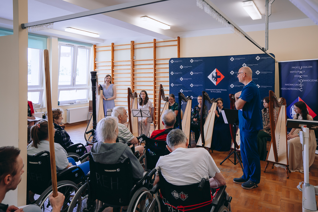 Na pierwszym planie odwróceni tyłem ludzie stanowiący publiczność, na drugim mężczyzna w niebieskim stroju, na trzecim siedem młodych kobiet stojących lub siedzących przy siedmiu harfach. Z tyłu drabinki gimnastyczne, ścianka reklamowa i roll-up.