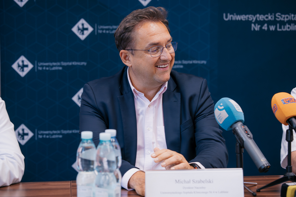 Uśmiechnięty mężczyzna w średnim wieku siedzący w garniturze przy stole konferencyjnym.  Przed nimi stoją mikrofony z logo różnych mediów i butelki wody. W tle widoczna jest granatowa ścianka reklamowa z logo Uniwersyteckiego Szpitala Klinicznego nr 4 w Lublinie.