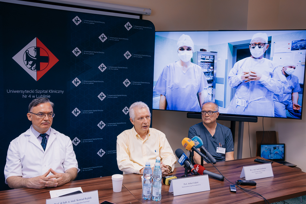 Na zdjęciu trzech mężczyzn siedzi przy stole konferencyjnym w sali. Po lewej stronie znajduje się prof. Konrad Rejdak w białym fartuchu lekarskim, pośrodku prof. Arthur Cukiert w żółtej koszuli, a po prawej dr n. med. Jacek Gawlewicz w granatowej odzieży medycznej. Przed nimi stoją mikrofony z logo różnych mediów. W tle widoczna jest granatowa ścianka reklamowa z logo Uniwersyteckiego Szpitala Klinicznego nr 4 w Lublinie oraz ekran ze zdjęciem przedstawiającym zespół operacyjny w sali zabiegowej – trzech lekarzy w maskach i strojach chirurgicznych.