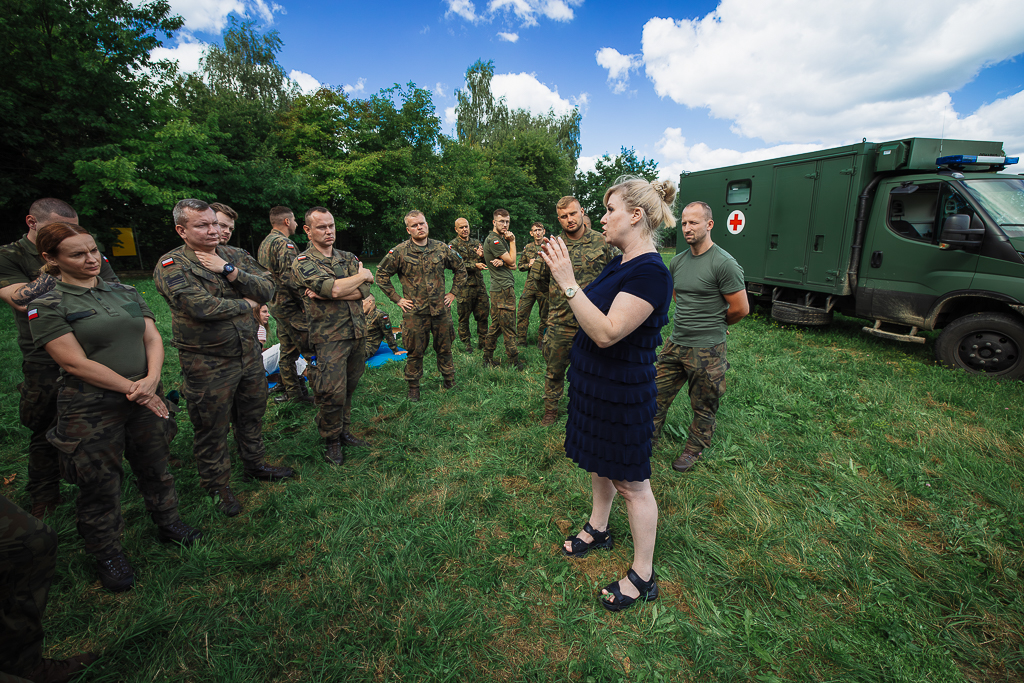 Blondynka w czarnej sukience mówi do osób w strojach wojskowych. Z tyłu wojskowy samochód.