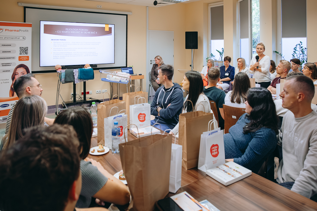 Na pierwszym planie siedzący przy stołach uczestnicy spotkania – kobiety z ciąży z partnerami. W głębi tablica, na której jest wyświetlany slajd dotyczący fizjologii porodu. Z lewej strony – pracownice Szpitala biorący udział w Dniach Otwartych oraz zaproszeni goście. Obok z mikrofonem w dłoni Anna Harasim-Raszkiewicz, opowiadająca zebranym o fizjologii porodu.