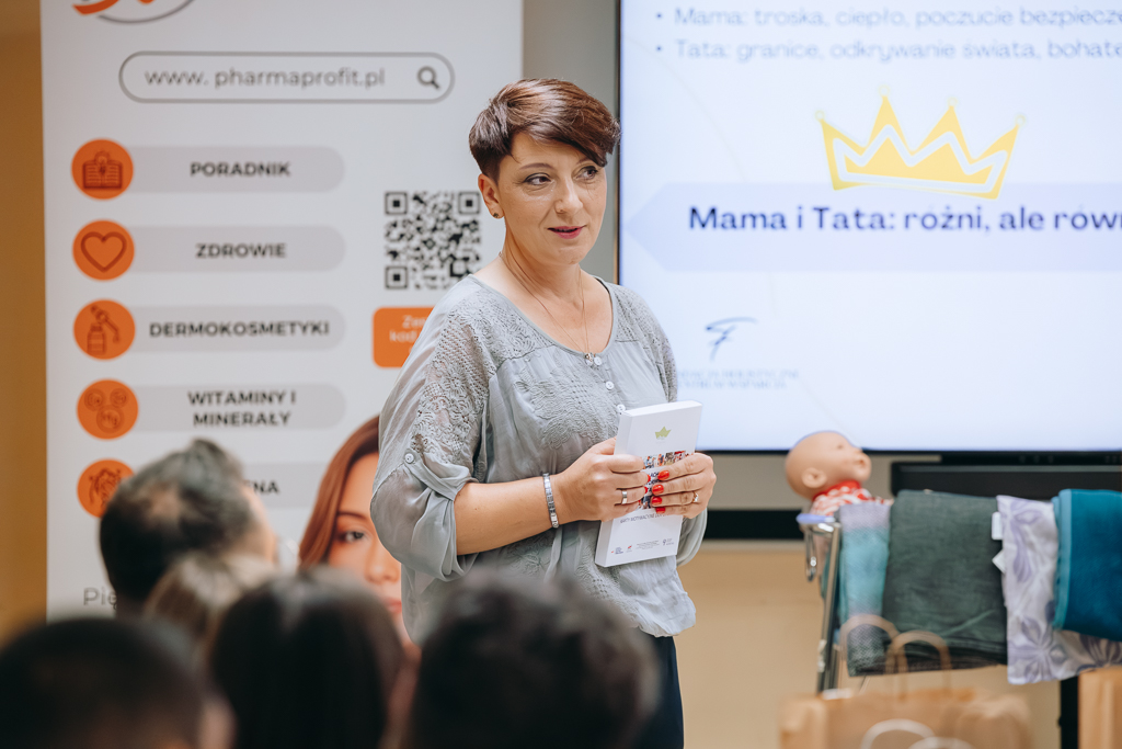 Prezes Fundacji „Holistyczne Centrum Wsparcia” Monika Krupa. W ręce trzyma pudełko z zestawem kart motywacyjnych dla ojców. W tle prezentowana przez Panią prezes prezentacja.