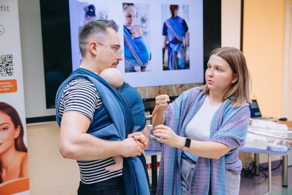Paulina Krzeszowska, prowadząca warsztaty z chustonoszenia prezentuje jednemu z uczestników spotkania sposób wiązania chusty. Za nimi widoczny slajd z prezentacji ze zdjęciami kobiety z założoną chustą do noszenia dziecka.