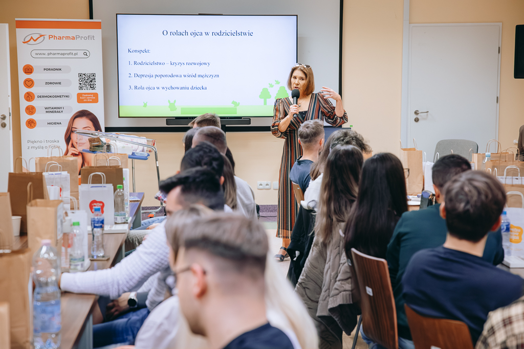 Dr Aneta Libera podczas swojej prezentacji, trzyma mikrofon w dłoni. Na pierwszym planie głowy uczestników spotkania. W tle prezentowana przez panią psycholog prezentacja.