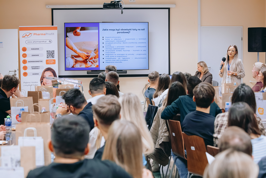 Siedzący przy stołach uczestnicy spotkania. Z lewej strony stoi opwiadająca o obowiązkach taty na sali porodowej położna Aleksandra Wrona. W tle wyświetlany jeden ze slajdów prezentacji do omawianego tematu.