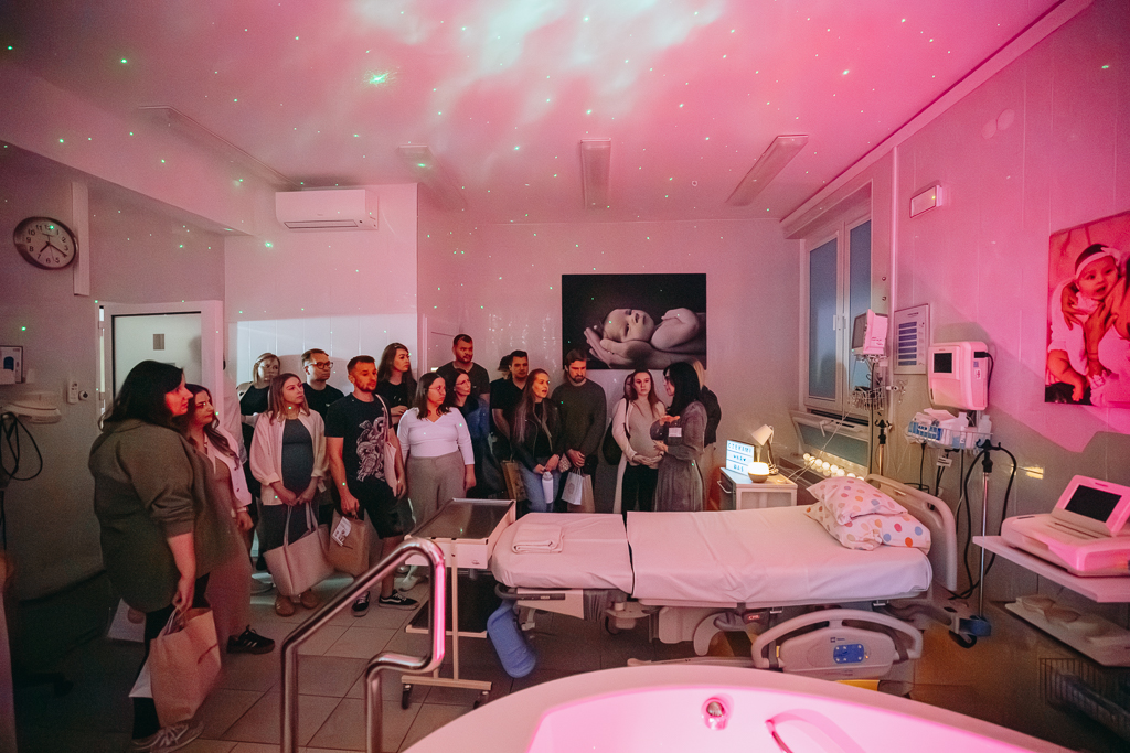 Uczestnicy spotkania podczas zwiedzania Traktu Porodowego. Są w sali porodowej z wanną, jej fragment jest widoczny na pierwszym planie. Obok szpitalne łóżko. W Sali jest też sprzęt medyczny. Na ścianek obrazki z noworodkami.