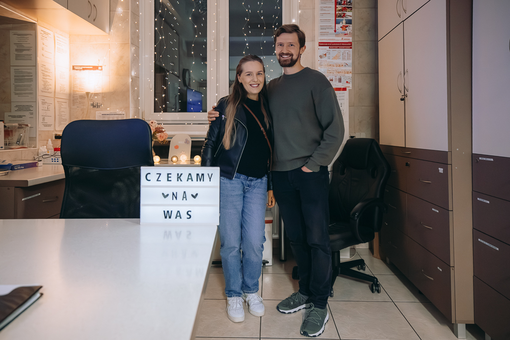 Uczestnicy spotkania - uśmiechnięci, stojący obok siebie, przytuleni pani Aleksandra i Pan Marek w dyżurce pielęgniarek. Obok wyposażenie - biurko oraz stojące wysokie szafy. Za parą klimatyczne drobne zapalone światełka. Na biurku podświetlany prostokątny napis: Czekamy na was.