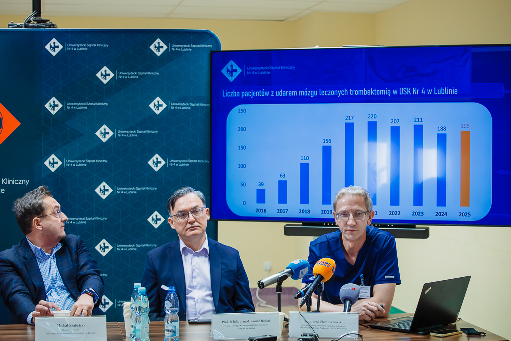 Trzech mężczyzn w średnim wieku przy stole z mikrofonami z tyłu monitor z wykresem i ścianka reklamowa z nazwą szpitala