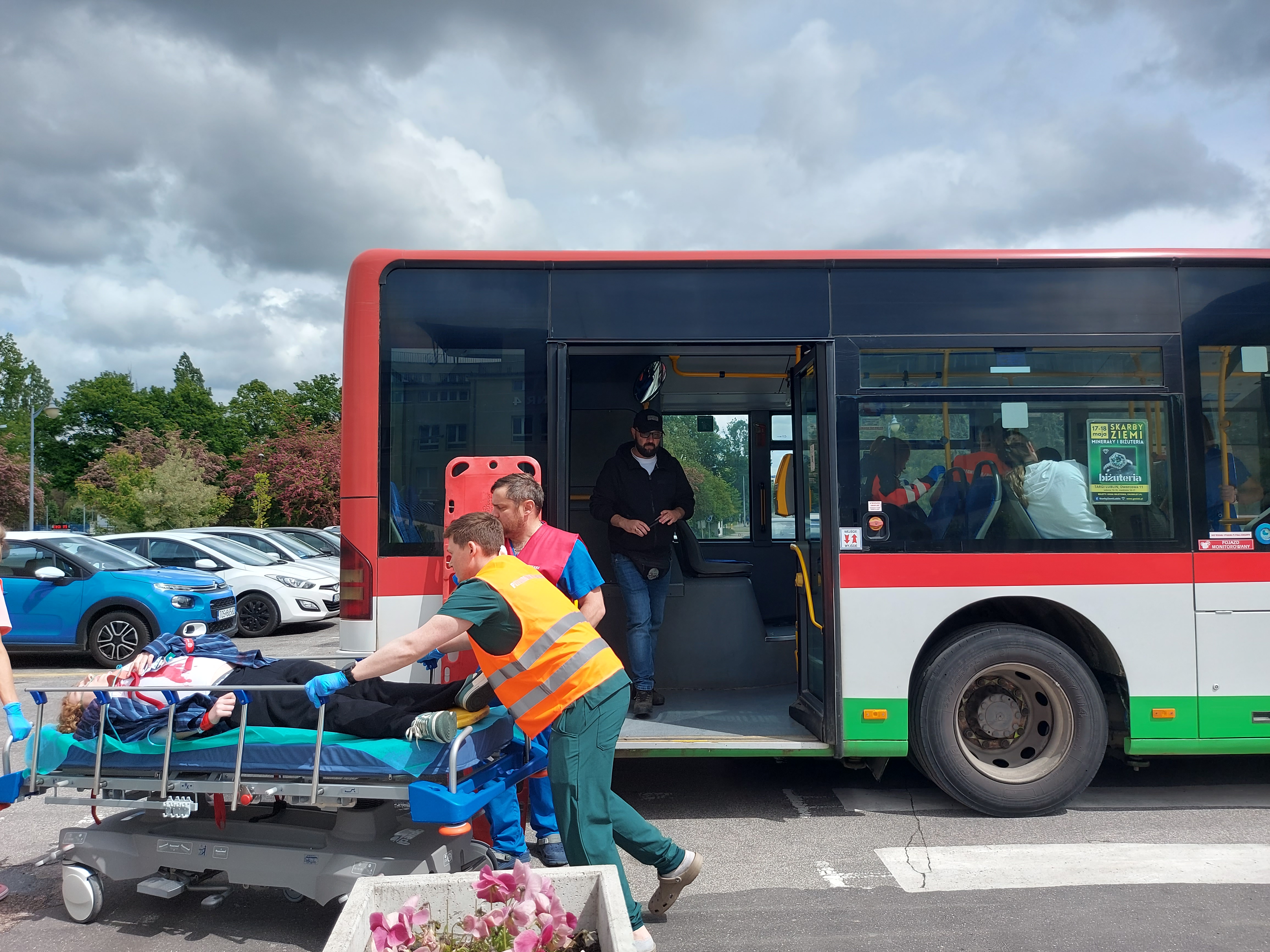 Tylnia część autobusu miejskiego z otwartymi drzwiami. Z przodu dwie osoby w kamizelkach odblaskowych pchają łóżko do przewożenia chorych na SOR. Na łóżku leży kobieta