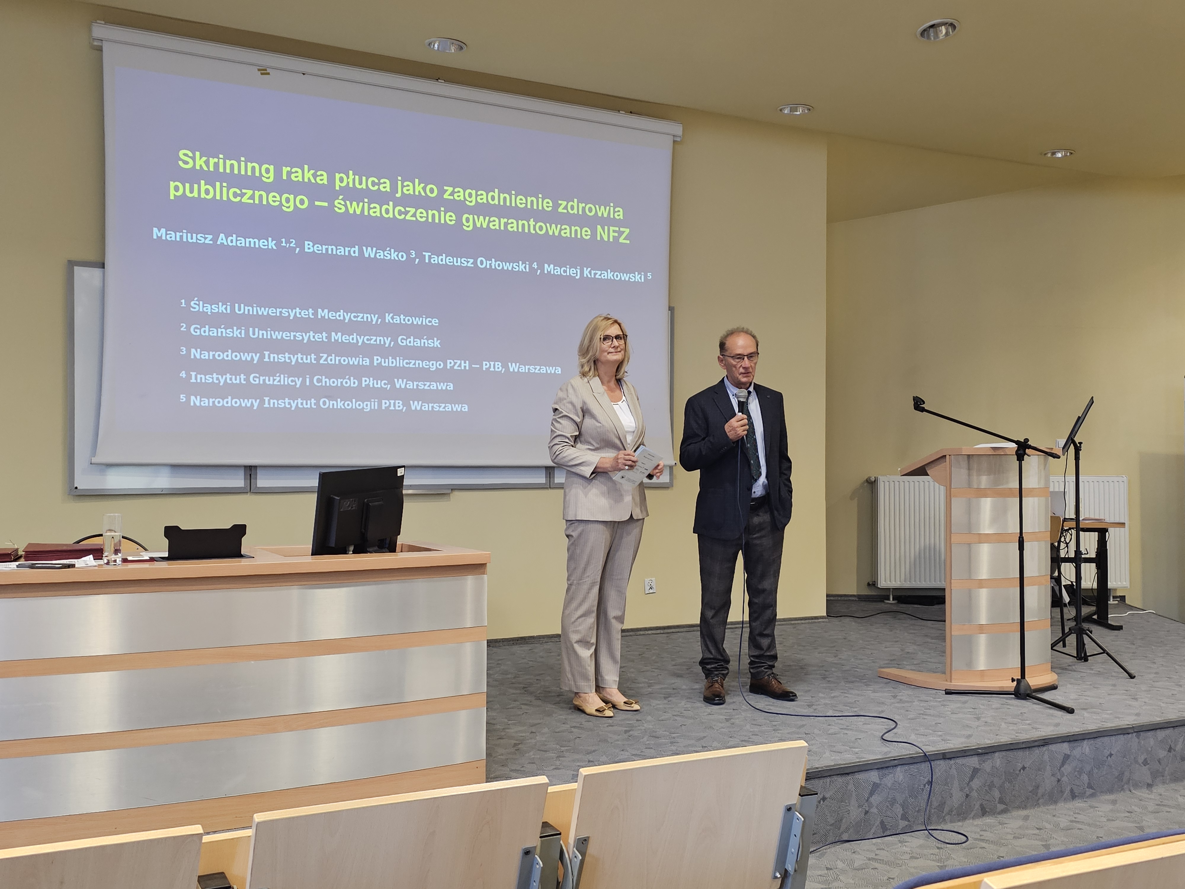 Elegancko ubrani kobieta i mężczyzna stojący na podwyższeniu w auli wykładowej, obok nich biurko i mównica. Mężczyzna trzyma mikrofon. Za nimi wyświetlony slajd prezentacji zaczynający się od słów: "Skrining raka płuca jako zagadnienie zdrowia publicznego" 