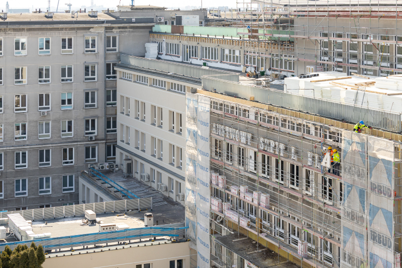 Widok z góry na wejście główne do szpitala, przy którym stoją rusztowania. Są na nich robotnicy demontujący stary napis. 