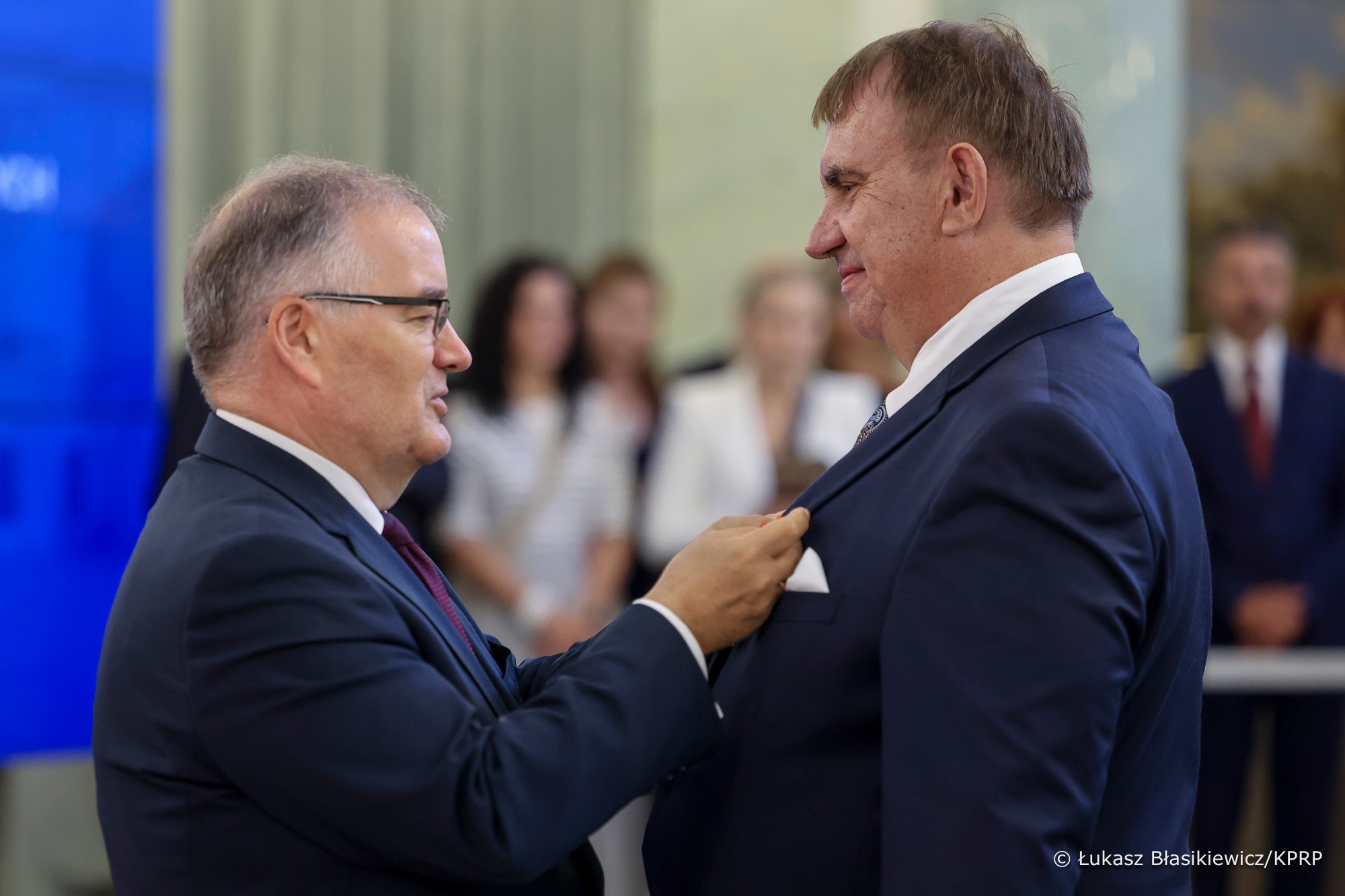 Dwóch mężczyzn w garniturach, jeden z dłonią przy klapie garnituru drugiego. Uśmiechają się. Z tyłu niewyraźne postacie.