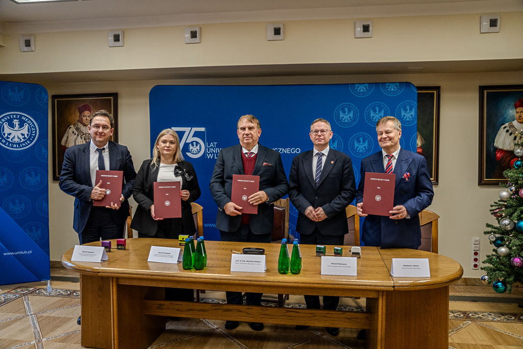 Sygnatariusze stoją za stołem w Sali Senatu Uniwersytetu Medycznego w Lublinie. Za nimi baner Uniwersytetu Medycznego w Lublinie. W dłoniach trzymają podpisane porozumienia w ozdobnej okładce.