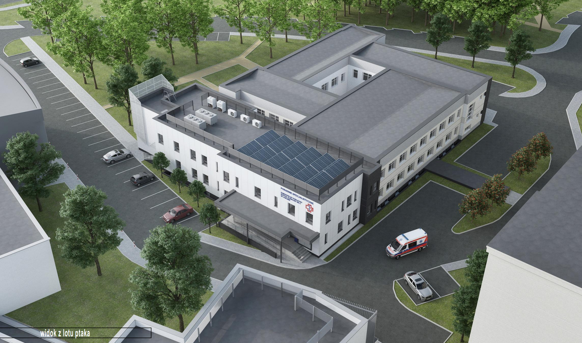 Na obrazie widoczny jest jasny, dwukondygnacyjny budynek oglądany z góry, z dziedzińcem w środku. Po lewej stronie fotografii znajduje się parking z kilkoma samochodami oraz fragment chodnika i drzew. Przy narożniku budynku widać ażurową, metalową osłonę oraz urządzenia ustawione na dachu. Na środkowej części dachu znajdują się panele słoneczne. Od frontu budynku widać wejście z zadaszeniem i schodami. Na ścianie umieszczony jest napis Poradnie Specjalistyczne oraz znak czerwonego krzyża. Po prawej stronie fotografii biegnie droga dojazdowa, po której jedzie karetka, a obok stoi zaparkowany samochód. Wokół budynku rosną drzewa, widać chodniki i fragmenty innych budynków. W lewym dolnym rogu obrazu znajduje się napis widok z lotu ptaka.
