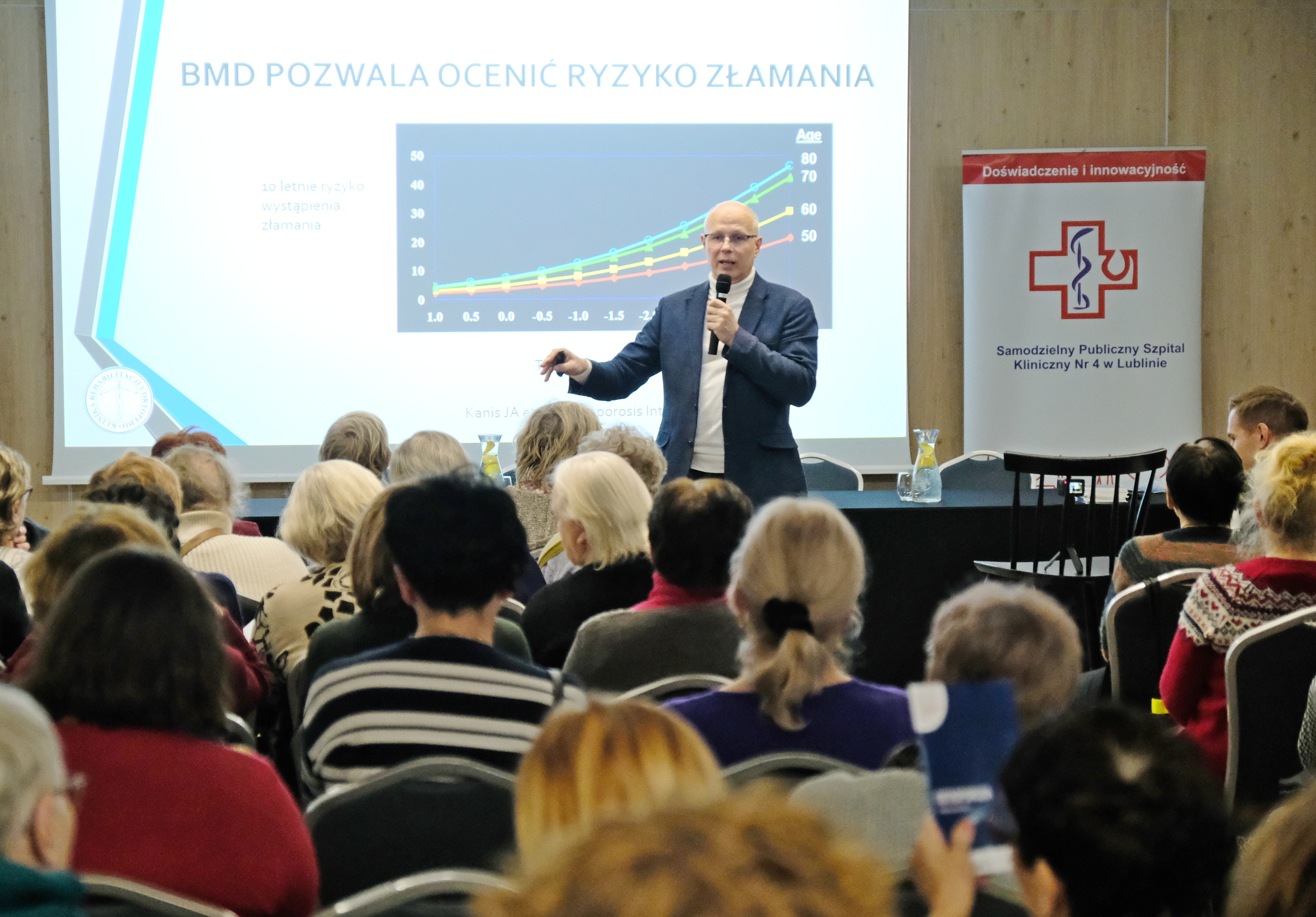 Mężczyzna przed prezentacją na ścianie i obok roll-upu z logo szpitala. Osoby na widowni widziane z tyłu.