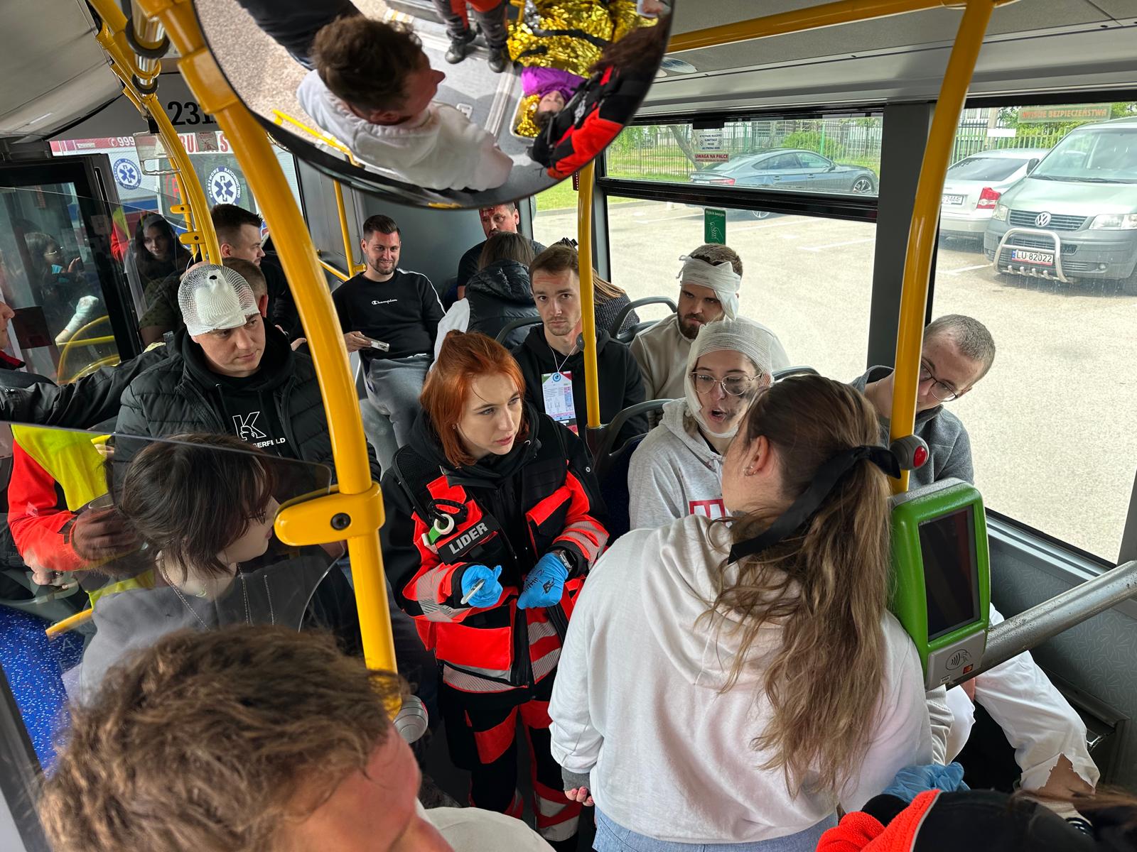 Środek autobusu miejskiego. Dużo ludzi, niektórzy w strojach służb ratowniczych, niektórzy z bandażami na głowach.