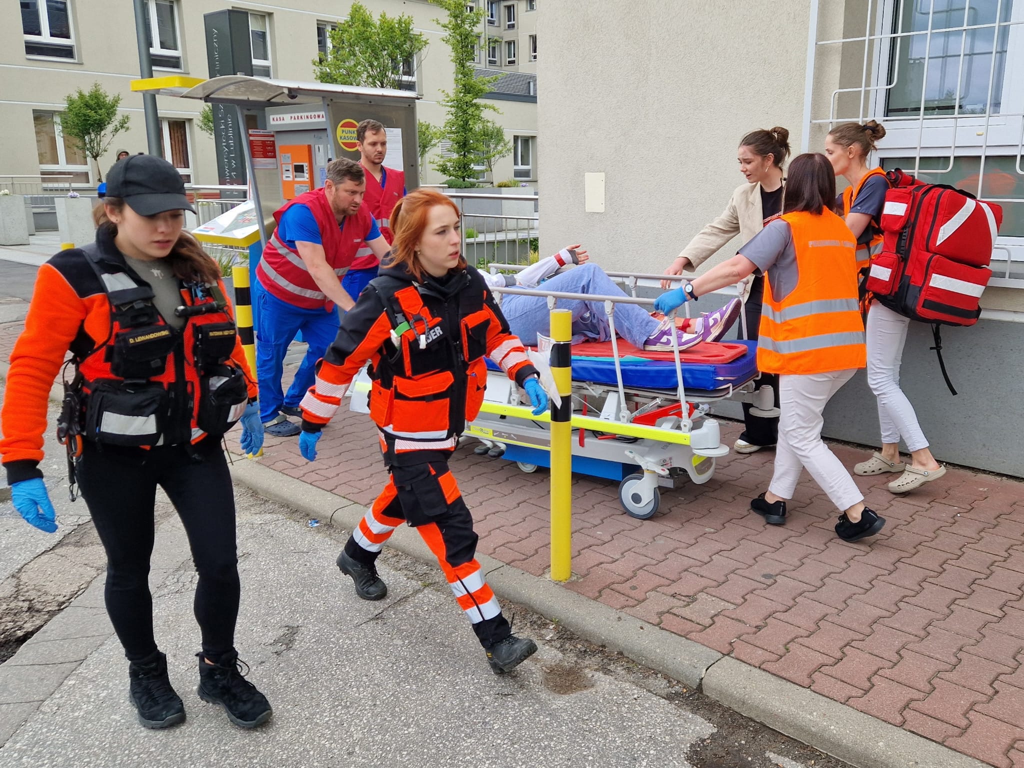 Sześć osób w strojach służb ratowniczych, jedna kobieta w marynarce, łóżko do transportu rannych, na którym leży siódma osoba.