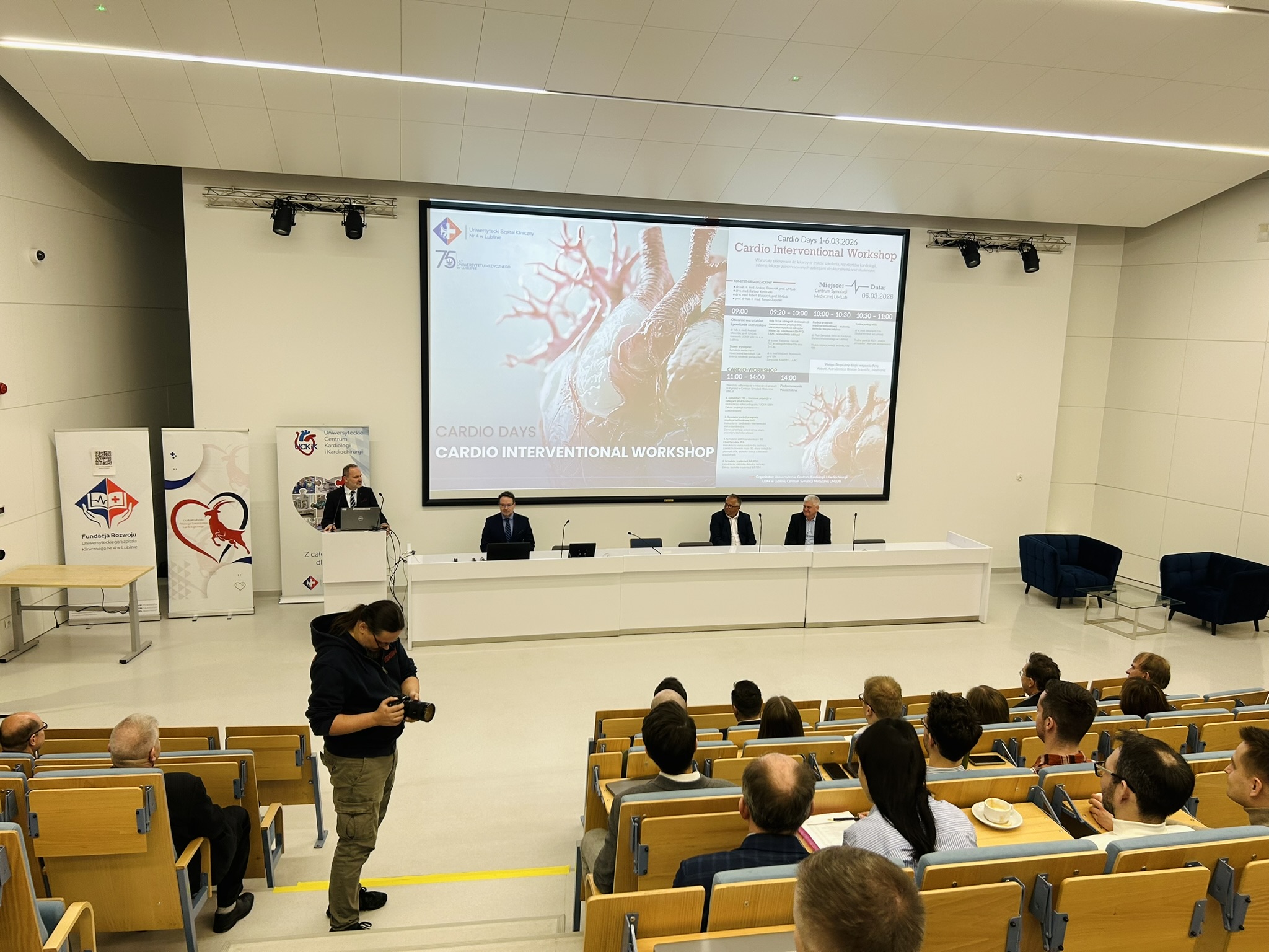 Na fotografii widać jasną salę wykładową. Po lewej stronie są stojące przy ścianie dwa pionowe banery z grafiką serca i logotypami instytucji. Obok znajduje się mównica, przy której stoi mężczyzna w garniturze i mówi do mikrofonu, patrząc w stronę publiczności. Na środku zdjęcia przed dużym białym stołem siedzi trzech mężczyzn, a za nimi na ścianie wisi bardzo duży ekran, na którym wyświetlony jest slajd z grafiką serca oraz programem wydarzenia. Przed sceną, nieco po lewej stronie, stoi mężczyzna trzymający w rękach aparat fotograficzny i patrzący w dół na jego ekran. W dolnej części zdjęcia oraz po prawej stronie widać rzędy drewnianych siedzeń ustawionych schodkowo, na których siedzą uczestnicy wydarzenia zwróceni w stronę sceny i ekranu. W tle po prawej stronie sali stoją dwa granatowe fotele i niewielki stolik, a cała przestrzeń jest jasna, z wysokim sufitem i liniowym oświetleniem.