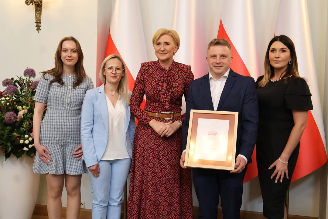 Cztery kobiety i jeden mężczyzna trzymający dyplom w ramce. Z tyłu flagi biało-czerwone.