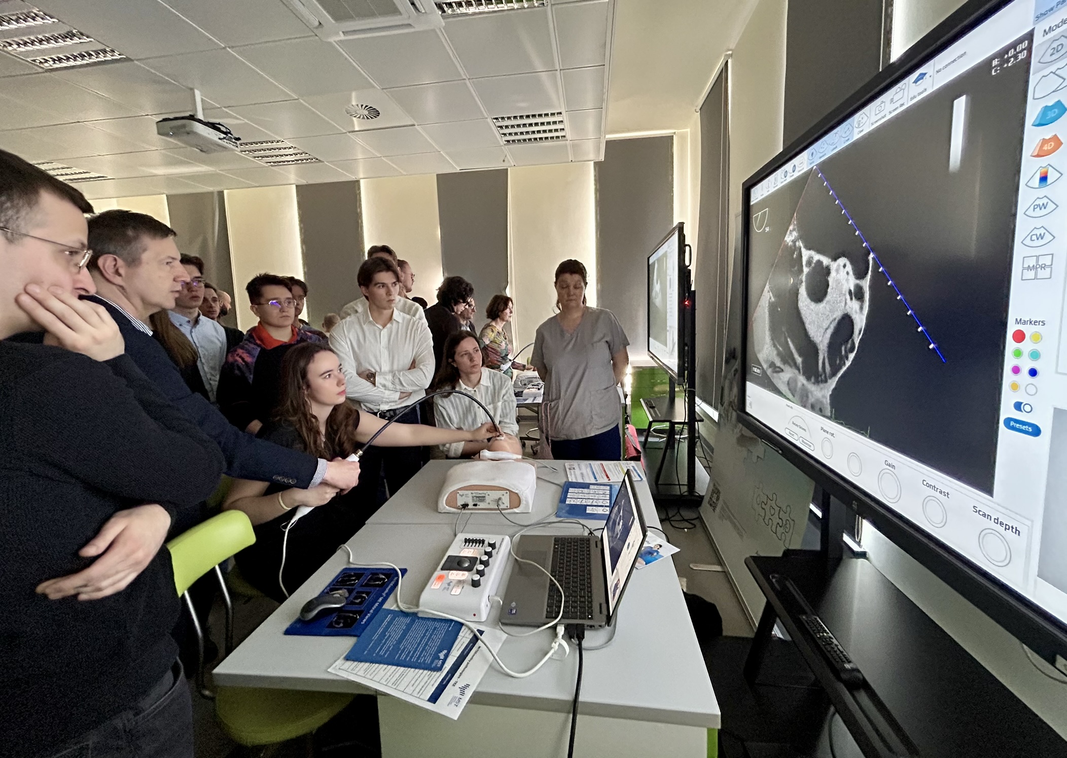 Po lewej stronie fotografii stoi kilka osób ustawionych blisko siebie i patrzących w prawą stronę, wśród nich widać młodych mężczyzn i kobiety, część z nich ma ręce skrzyżowane lub oparte na twarzy. Na środku zdjęcia przy stole siedzi młoda kobieta, która trzyma w dłoni długą sondę połączoną przewodem z urządzeniem stojącym na stole, a obok niej leży model części ludzkiego ciała oraz komputer przenośny i inne niewielkie urządzenia z kablami. Za stołem stoi kobieta w jasnym stroju i również patrzy w stronę dużego ekranu. Po prawej stronie fotografii znajduje się duży monitor ustawiony pionowo na stojaku, na którym wyświetlony jest czarno biały obraz wnętrza ludzkiego ciała oraz różne kolorowe ikony i przyciski. W tle widać jasną salę z wysokim sufitem, lampami, ekranem do wyświetlania obrazu oraz kilkoma osobami stojącymi dalej przy innych stanowiskach.