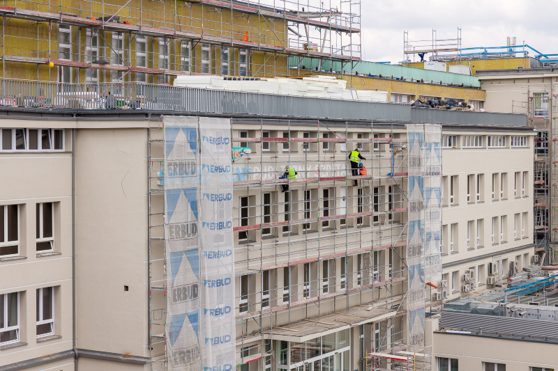 Widok na wejście główne do szpitala. Na rusztowaniu pracują osoby montujące nowy napis. 
