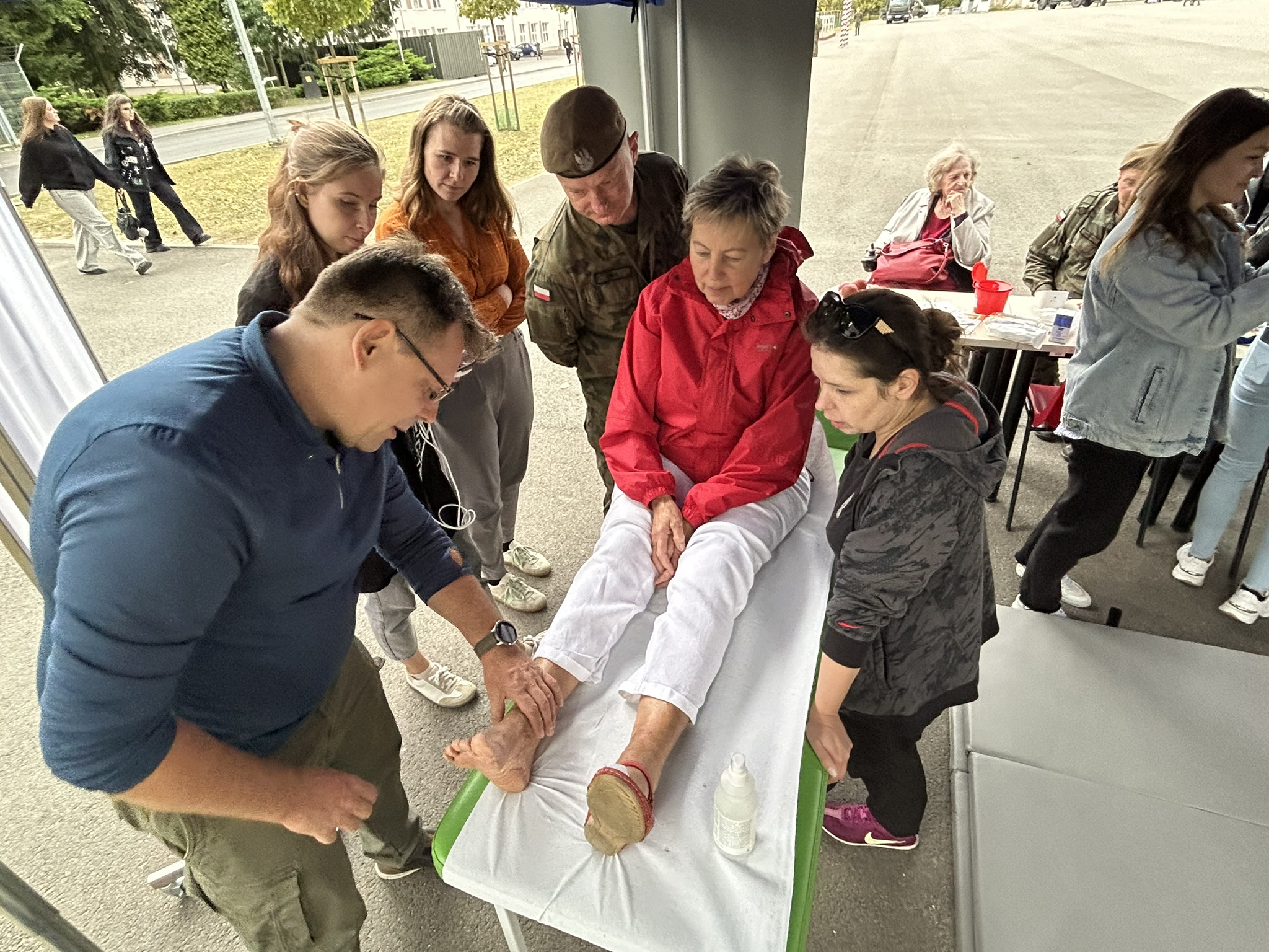 Na zdjęciu widać grupę osób zgromadzonych wokół starszej kobiety siedzącej na leżance. Ma na sobie czerwoną kurtkę i białe spodnie, a jej nogi są wyciągnięte przed siebie. Mężczyzna w okularach, stojący obok, dotyka jej stopy i uważnie się jej przygląda. Obok niego stoi młoda kobieta, która również obserwuje sytuację. Za nimi stoi kilkuosobowa grupa, w tym mężczyzna w mundurze wojskowym i dwie młode kobiety. W tle widać kilka osób siedzących przy stole i przechodniów. Atmosfera jest spokojna i skupiona.