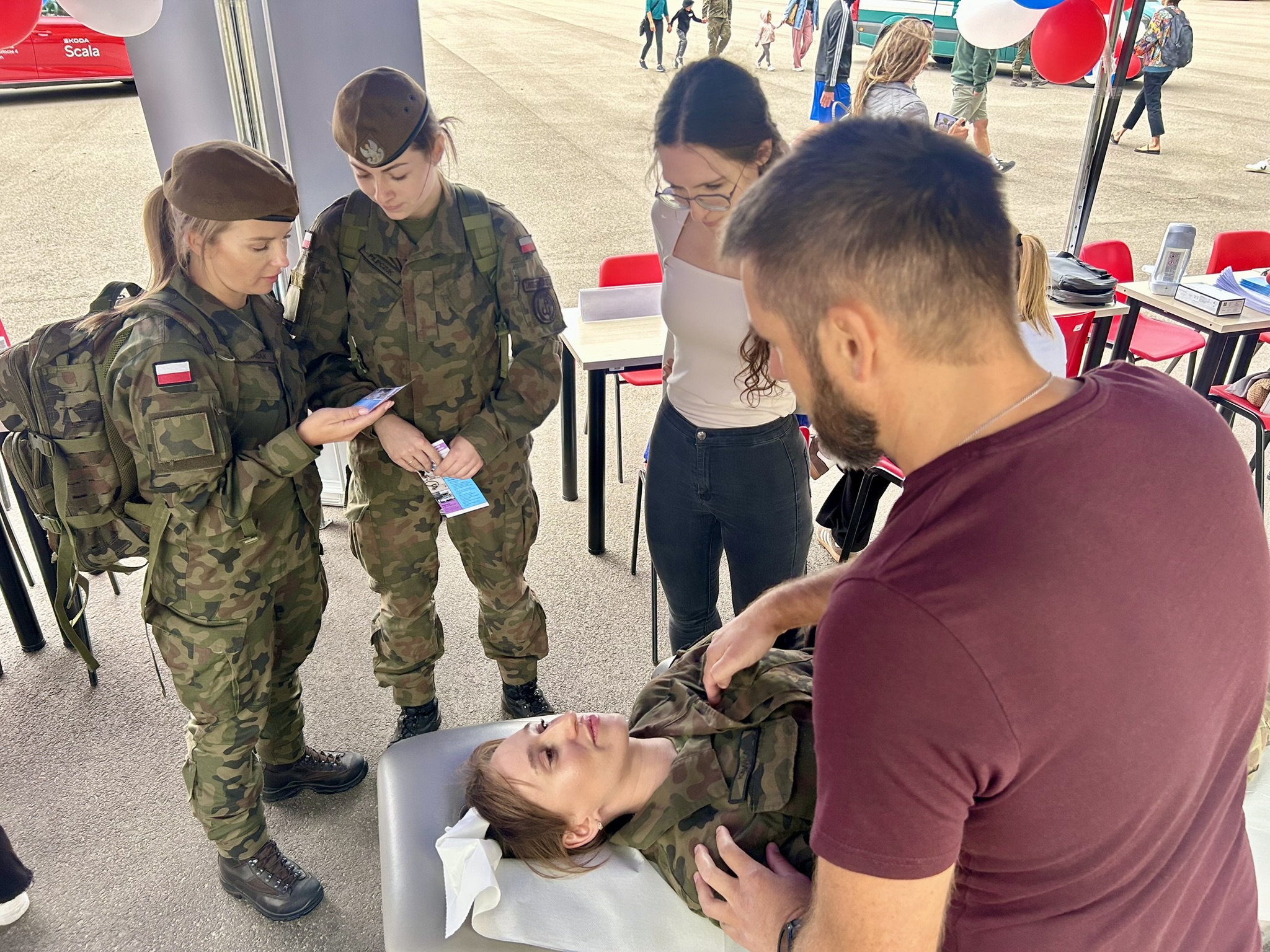 Na zdjęciu widać cztery kobiety i mężczyznę. Trzy kobiety są w strojach wojskowych, jedna z nich leży na leżance twarzą do góry, a na jej barkach są ręce mężczyzny, reszta kobiet przygląda się temu procesowi.
