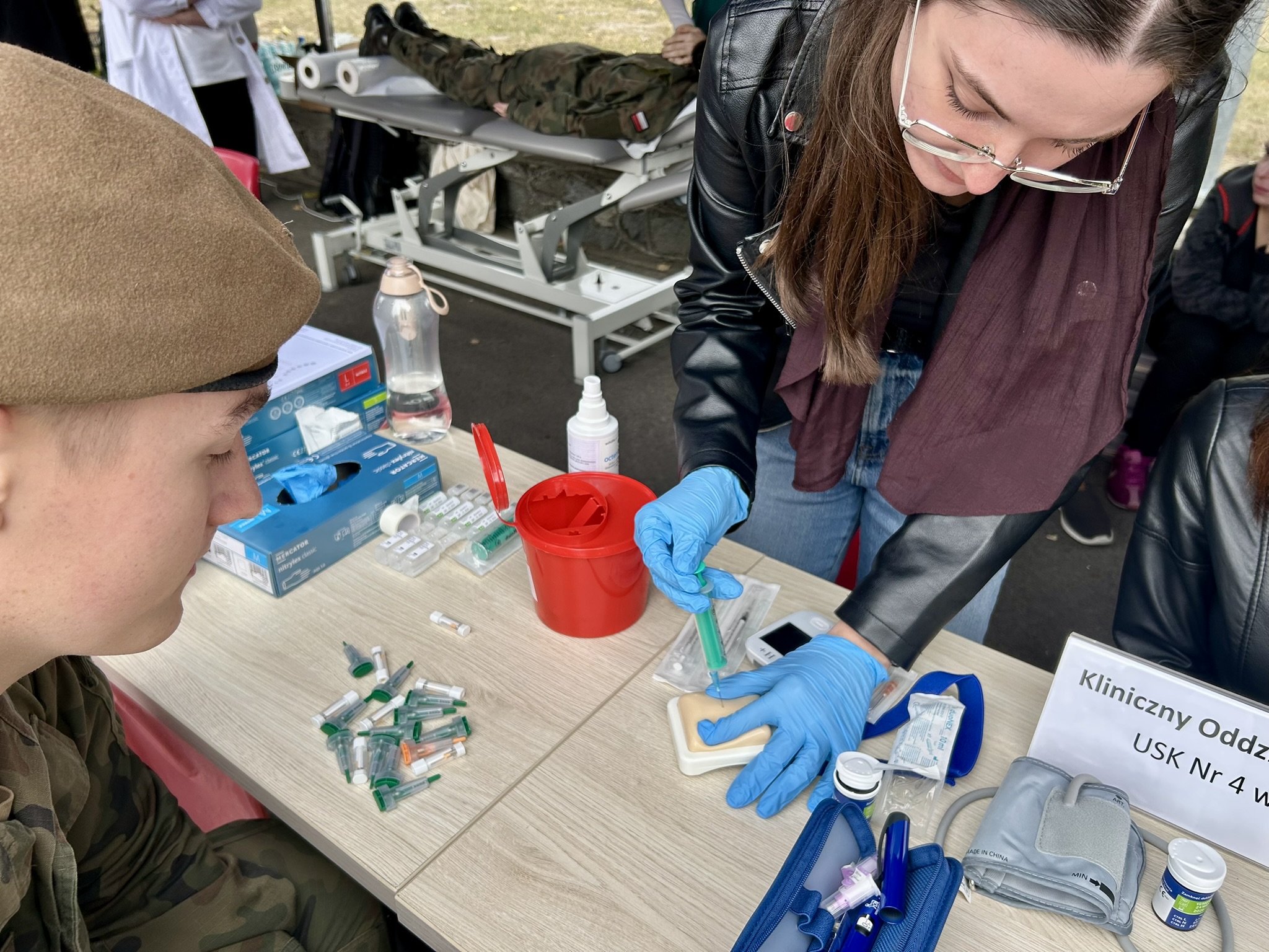 Na zdjęciu widać dwa połączone ze sobą stoły lub biurka, po jednej stronie młoda kobieta w okularach i rękawiczkach medycznych wykonuje prezentację procedury przy użyciu strzykawki, czemu przygląda się młody mężczyzna w berecie i stroju wojskowym.