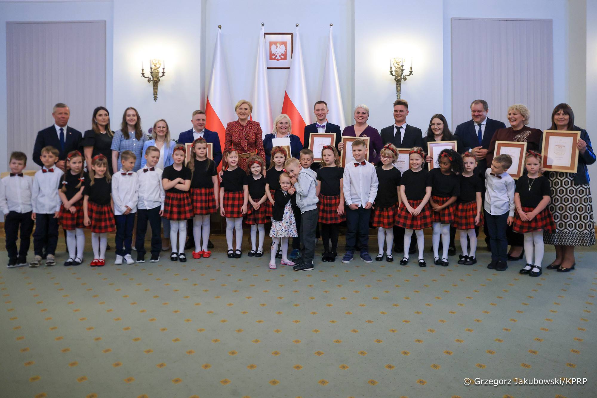 Wiele osób ustawionych w dwóch rzędach, w pierwszym rzędzie dzieci. Wszyscy ubrani elegancko. Z tyłu biało-czerwone flagi i godło RP.