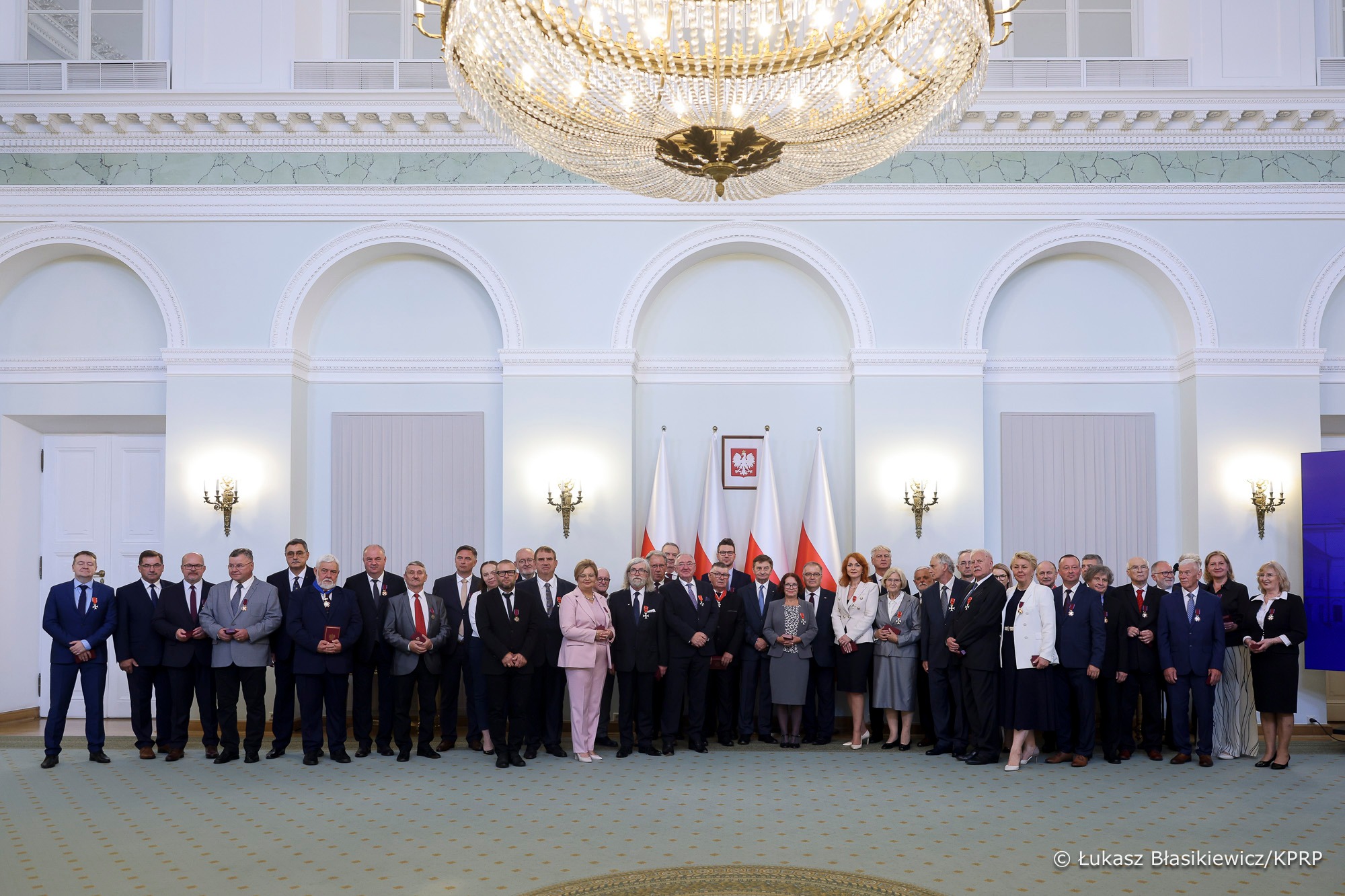 Zdjęcie przedstawia uroczyste, grupowe zdjęcie wykonane w eleganckiej sali z dużym, bogato zdobionym żyrandolem zwisającym z sufitu. W tle widać jasnoniebieskie ściany z klasycznymi zdobieniami i łukowatymi wnękami oraz polskie flagi i godło Rzeczypospolitej Polskiej umieszczone centralnie. Przed flagami stoi liczna grupa osób – kobiety i mężczyźni w różnym wieku, ubrani w odświętne stroje: garnitury, marynarki, garsonki i sukienki. Większość z nich trzyma w dłoniach pudełka z medalami lub ma odznaczenia przypięte do piersi.
