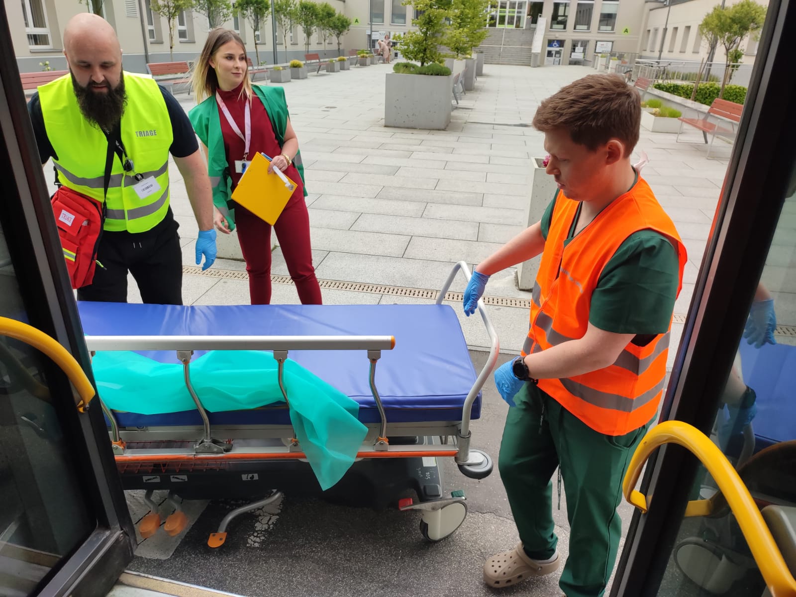 Dwóch mężczyzn w odblaskowych kamizelkach, kobieta trzymająca teczkę obok pustego łóżka do transportu rannych. Na pierwszym planie otwarte drzwi autobusu miejskiego