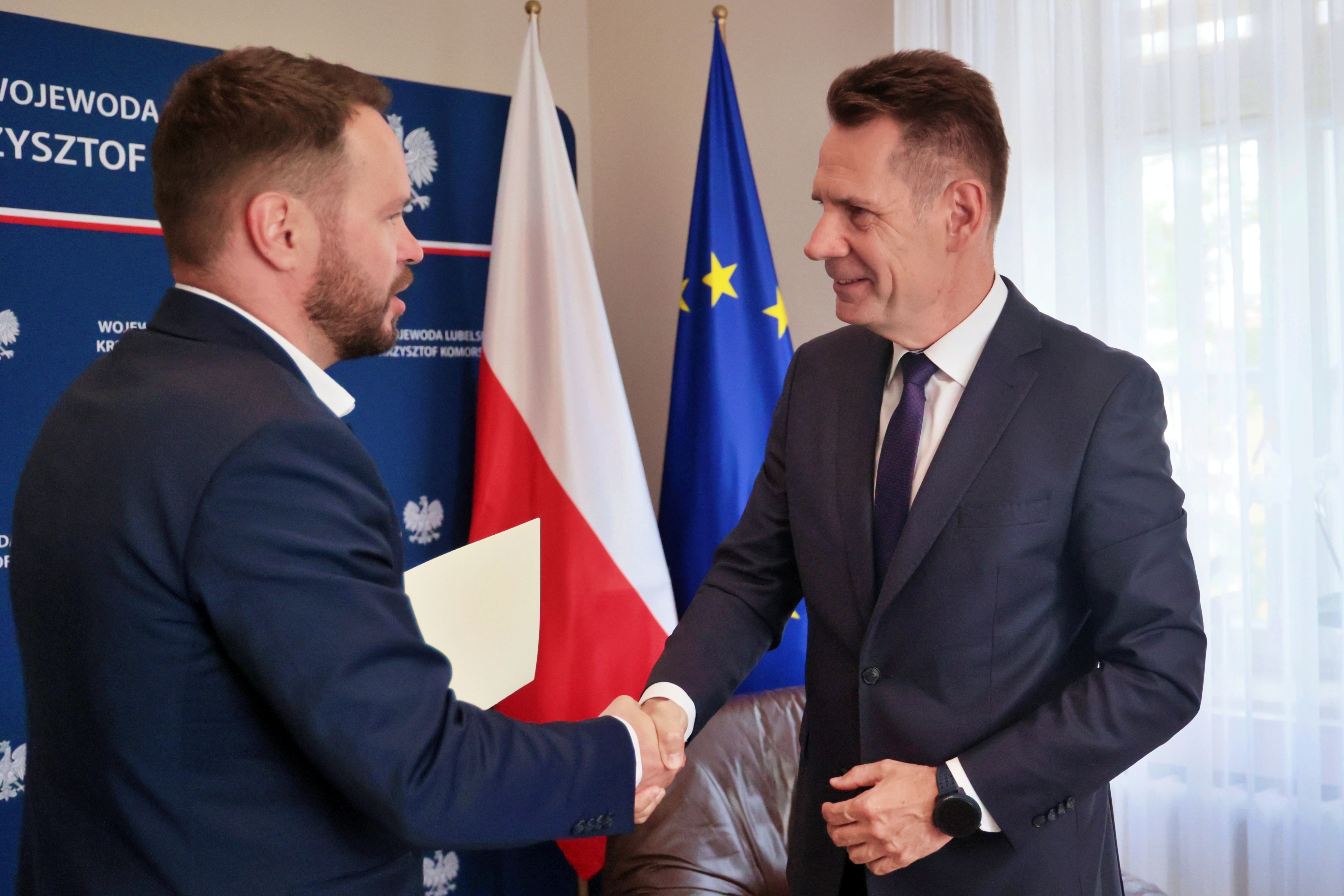 Elegancko ubrany mężczyzna ściska dłoń elegancko ubranemu mężczyźnie. Za nimi flagi Polski i Unii Europejskiej.