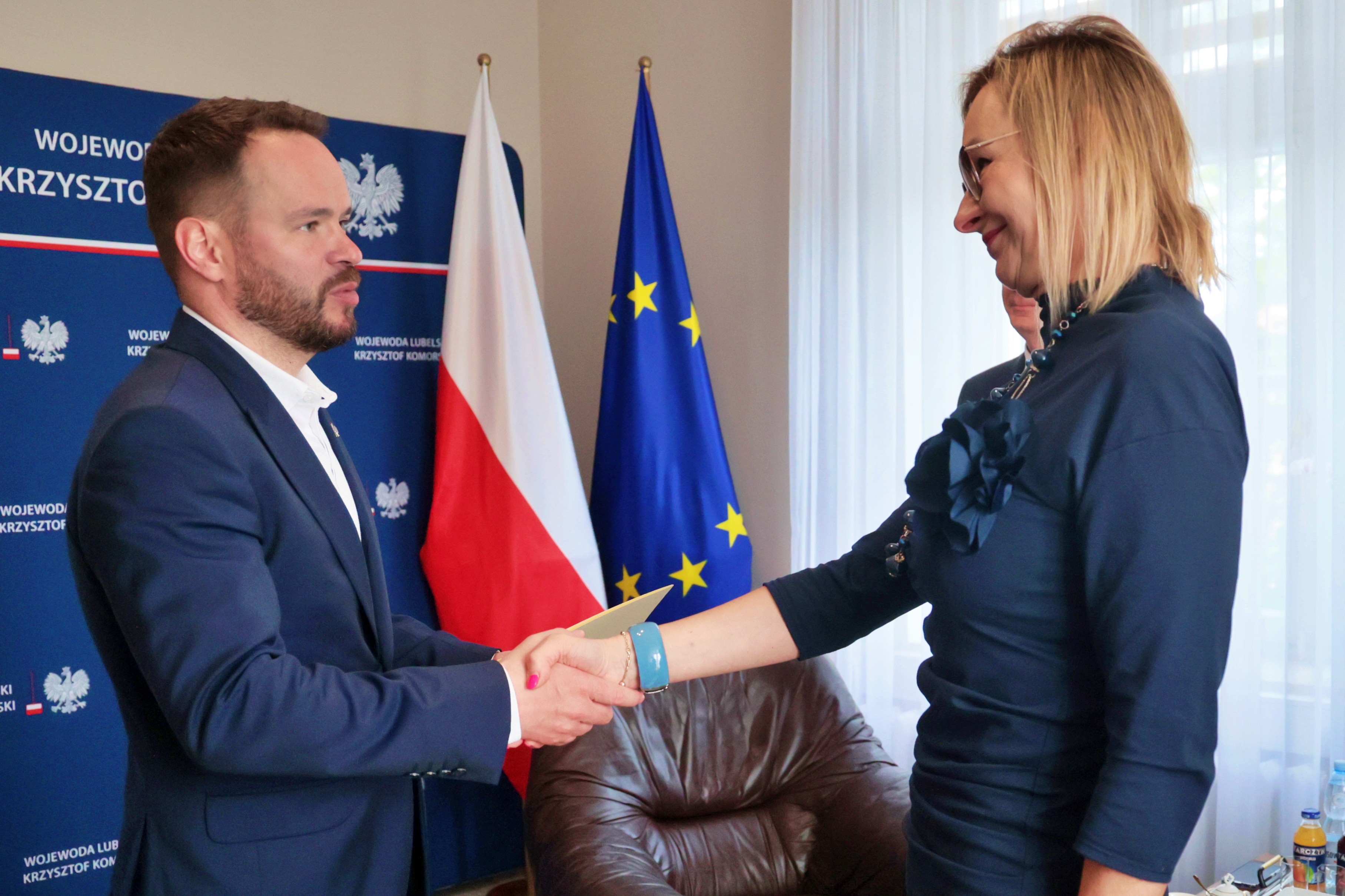 Elegancko ubrany mężczyzna ściska dłoń elegancko ubranej kobiecie. Za nimi flagi Polski i Unii Europejskiej.