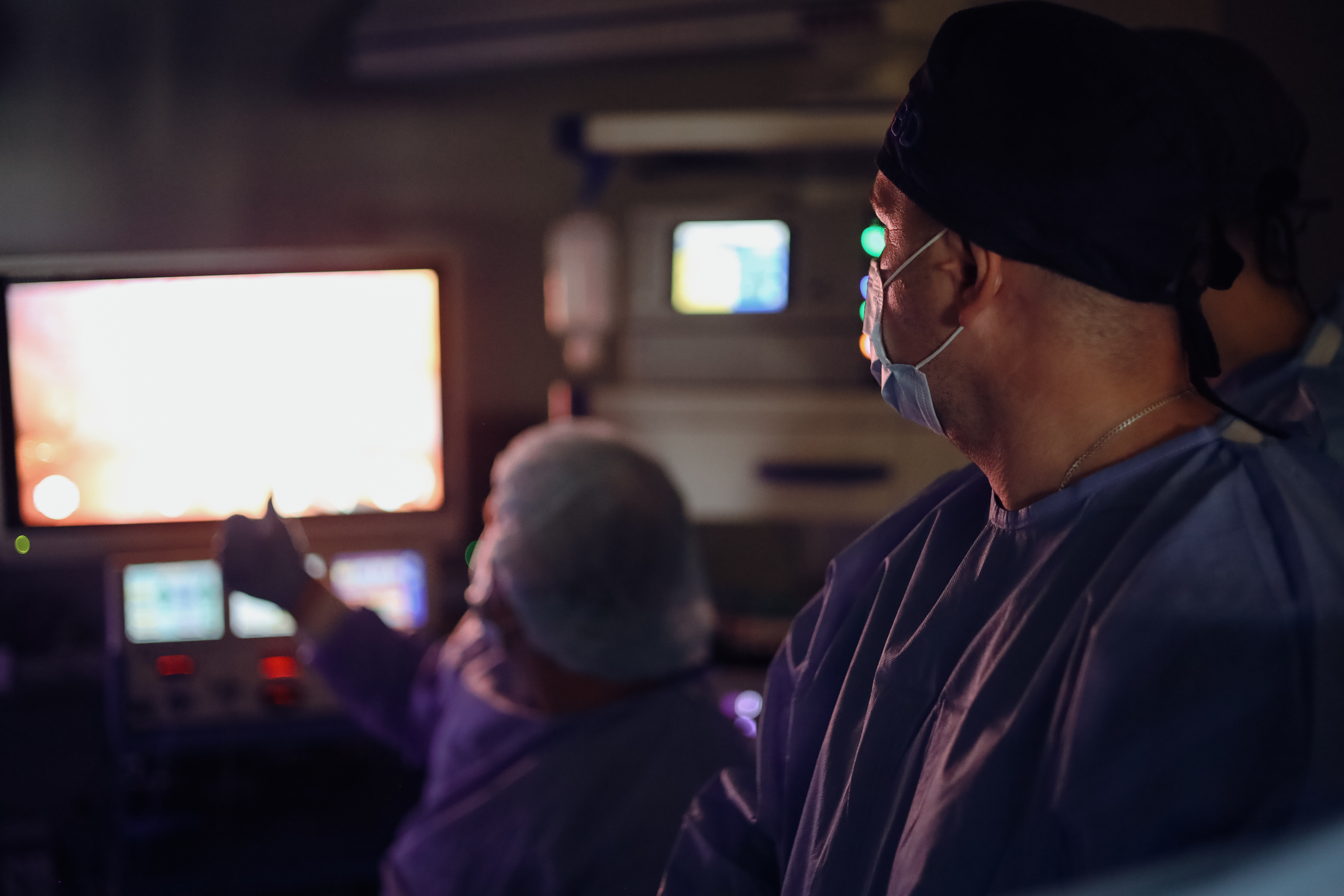 Na zdjęciu widać dwie osoby w niebieskich strojach ochronnych i maseczkach. Jedna z nich siedzi i wskazuje coś na dużym, jasno świecącym ekranie, druga stoi obok i również patrzy w jego stronę. W tle widoczne są urządzenia z kolorowymi światełkami i monitorami. Całość sprawia wrażenie skupionej pracy przy nowoczesnym sprzęcie.