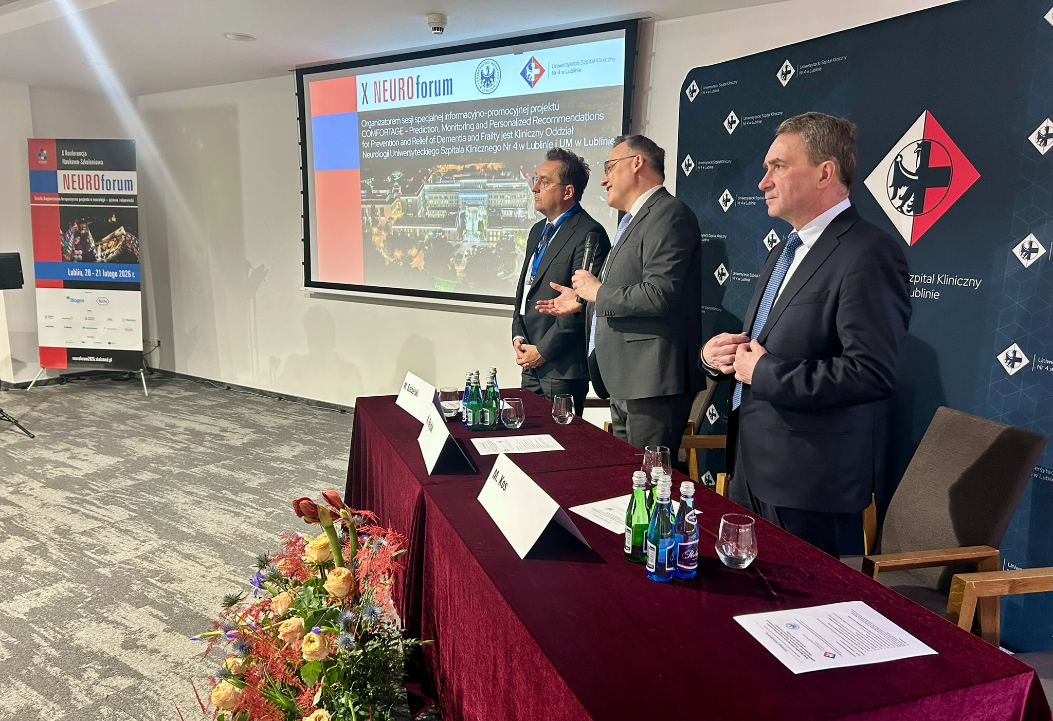 Na zdjęciu widać trzech mężczyzn stojących za długim stołem przykrytym bordowym obrusem w sali konferencyjnej. Po lewej stronie fotografii stoi rollup z napisem neuroforum oraz informacją o miejscu i dacie wydarzenia, a obok na ścianie wisi duży ekran z wyświetloną prezentacją zawierającą tytuł x neuroforum, logotypy oraz zdjęcie miasta nocą. Na środku zdjęcia, przed stołem, stoi kompozycja kwiatowa z jasnymi i czerwonymi kwiatami oraz zielonymi liśćmi. Na stole ustawione są szklanki, butelki z wodą oraz kartki z nazwiskami. Po lewej stronie spośród trzech mężczyzn stoi osoba w ciemnym garniturze, białej koszuli i krawacie, ma okulary oraz identyfikator na szyi. W centrum stoi mężczyzna w szarym garniturze i jasnym krawacie, trzyma w ręce mikrofon i gestykuluje drugą dłonią. Po prawej stronie fotografii stoi mężczyzna w ciemnym garniturze i niebieskim krawacie, ma ręce złożone przed sobą. Tło po prawej stronie stanowi ścianka z powtarzającym się logo uniwersyteckiego szpitala klinicznego nr 4 w lublinie. Podłoga jest pokryta jasną wykładziną, a oświetlenie jest jasne i równomierne.