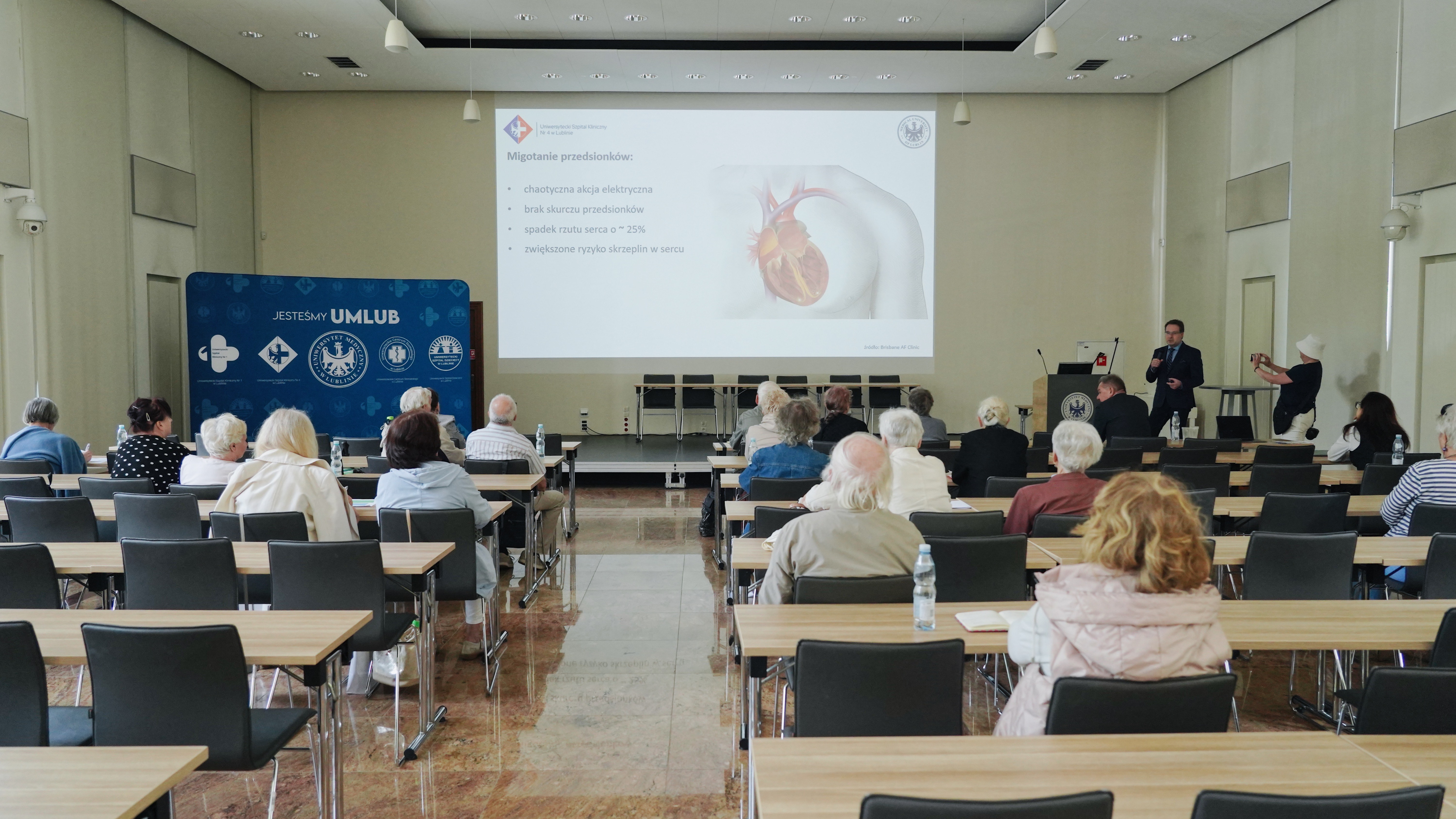 Kilkadziesiąt starszych osób odwróconych plecami, siedzących w ławkach. Z tyłu mężczyzna w garniturze trzymający mikrofon, kobieta w białej czapce robiąca mu zdjęcie. Na ścianie wyświetlona prezentacja z widocznym sercem człowieka. Z boku ścianka z napisem "jesteśmy UMLub" i logami Uniwersytetu Medycznego w Lublinie i lubelskich szpitali klinicznych.