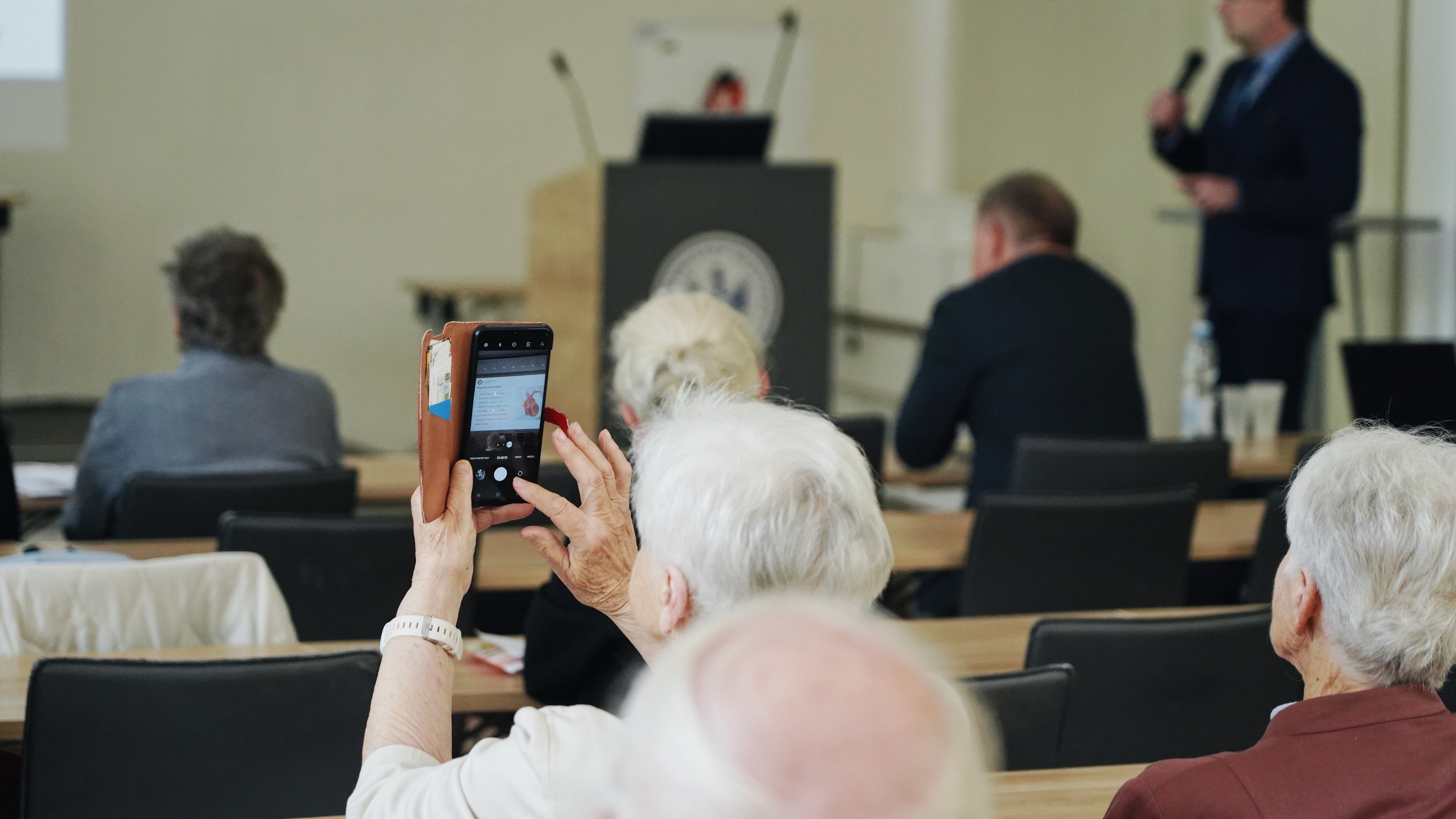 Starsze osoby siedzące za stołami, odwrócone plecami, jedna trzyma wysoko telefon robiąc zdjęcie. Z przodu widać osobę mówiącą do mikrofonu. To mężczyzna w średnim wieku.