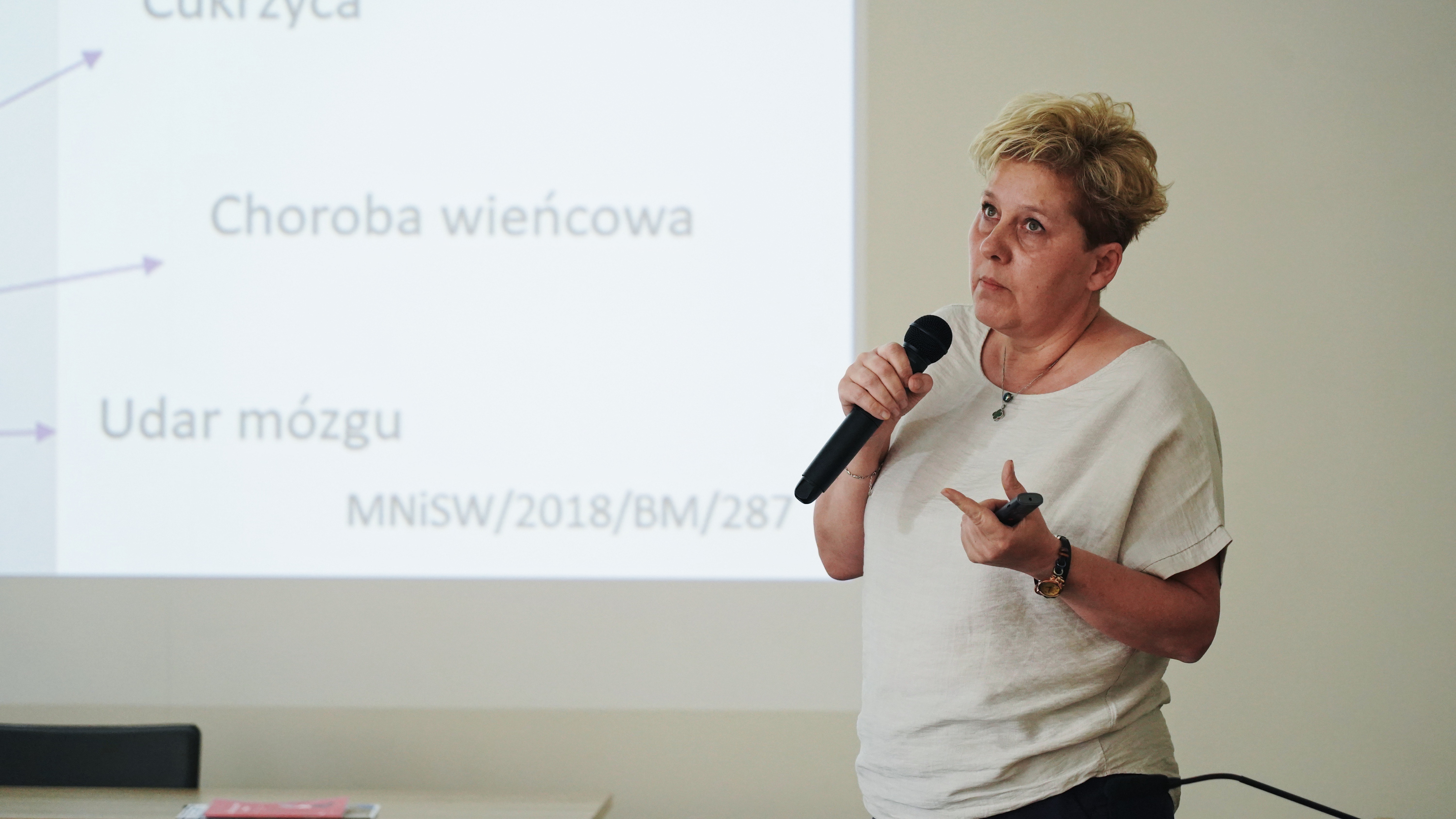 Kobieta w średnim wieku, w jasnej bluzce, mówiąca do mikrofonu. Za nią widoczna część prezentacji z rzutnika, na której widać słowa "choroba wieńcowa, udar mózgu, cukrzyca"