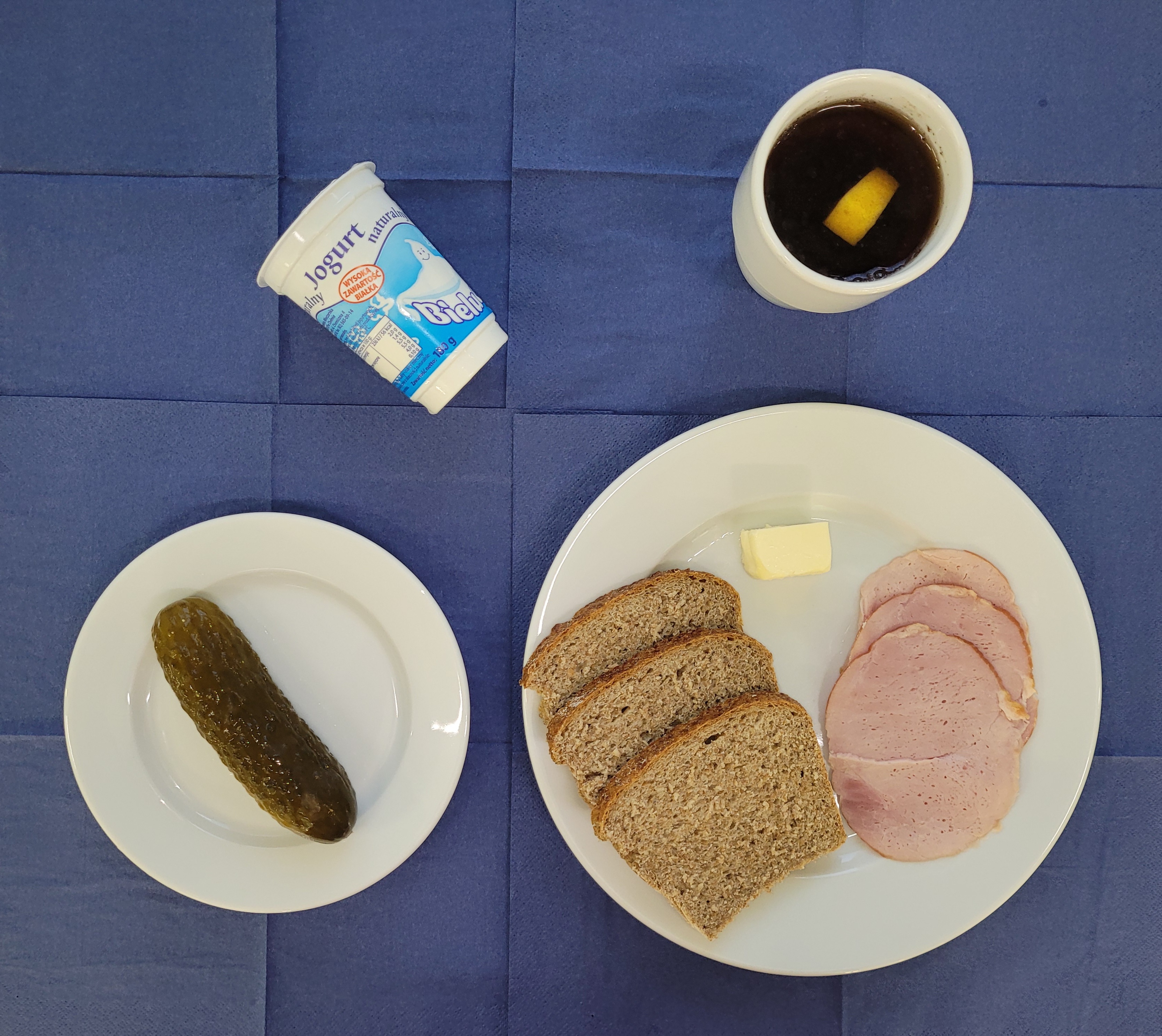 na talerzach chleb razowy, masło, szynka gotowana, ogórek kiszony, jogurt naturalny, w kubku herbata z cytryną gorzka